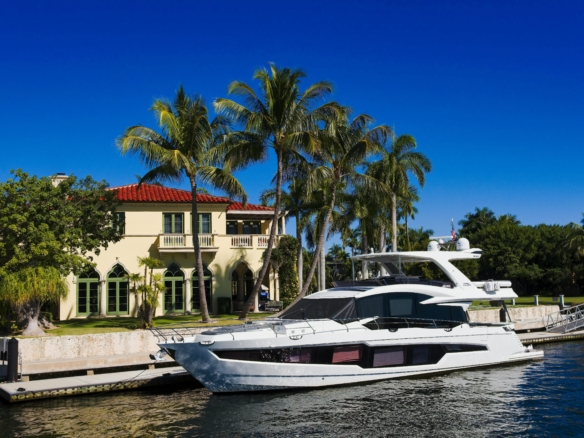 Luxury yacht docked by a waterfront mansion with palm trees under a clear blue sky.