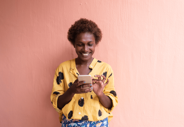 Smiling woman in a yellow sweater using a smartphone against a peach background.