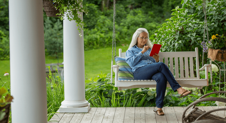 Elderly woman reading a book on a porch swing surrounded by greenery.