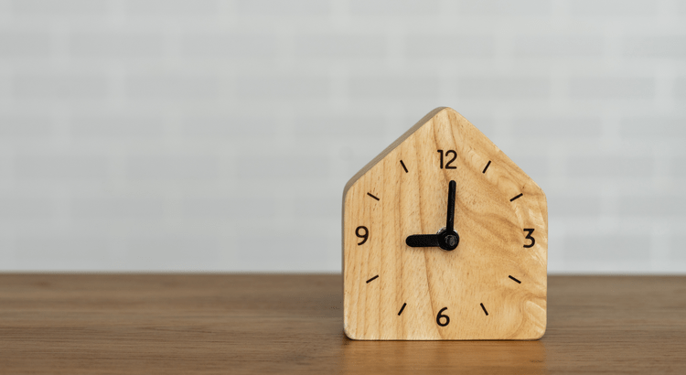 Wooden clock shaped like a house showing 3 o'clock on a wooden surface.